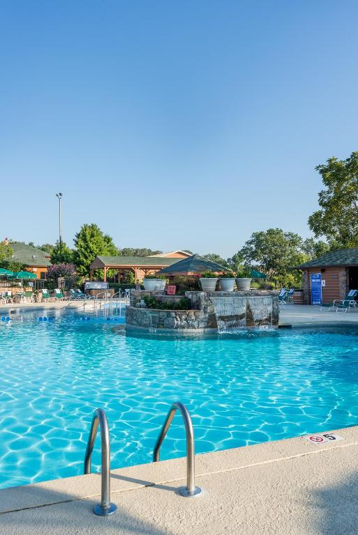 Outdoor swimming pool with clear blue water, surrounded by lounge chairs and trees under a clear sky. Stone feature in the center with plants on top. Ladder in the foreground.