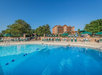 Outdoor pool area with clear blue water, surrounded by lounge chairs and green umbrellas. A large building and trees are in the background under a clear blue sky.