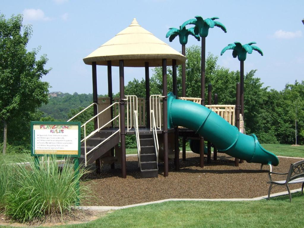 A playground with a slide, palm tree decorations, and a safety rules sign. Nearby are a bench and grassy areas.
