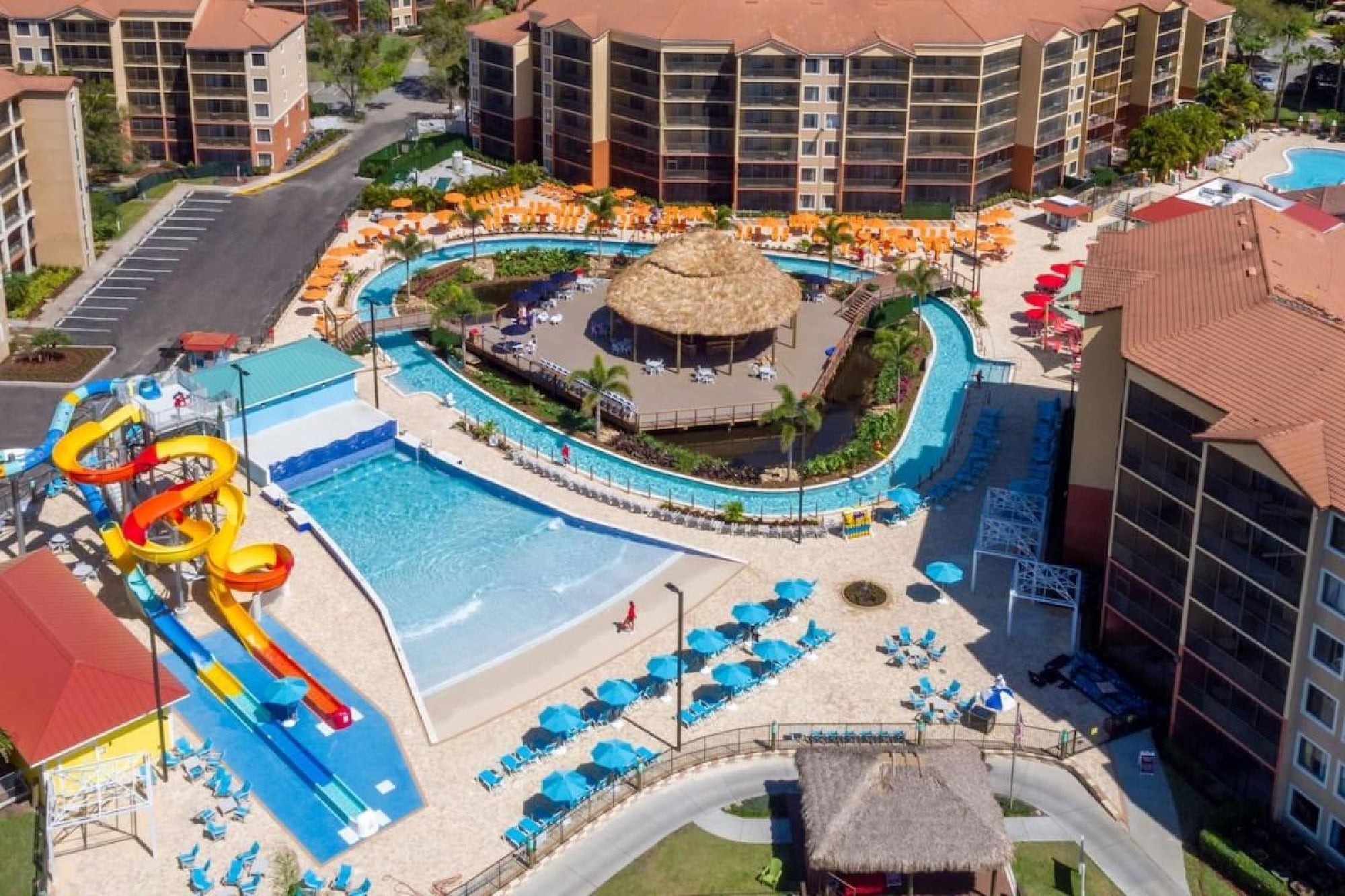 Aerial view of a resort featuring a water park with colorful slides, a lazy river, lounge chairs, and surrounding multi-story buildings with red roofs.