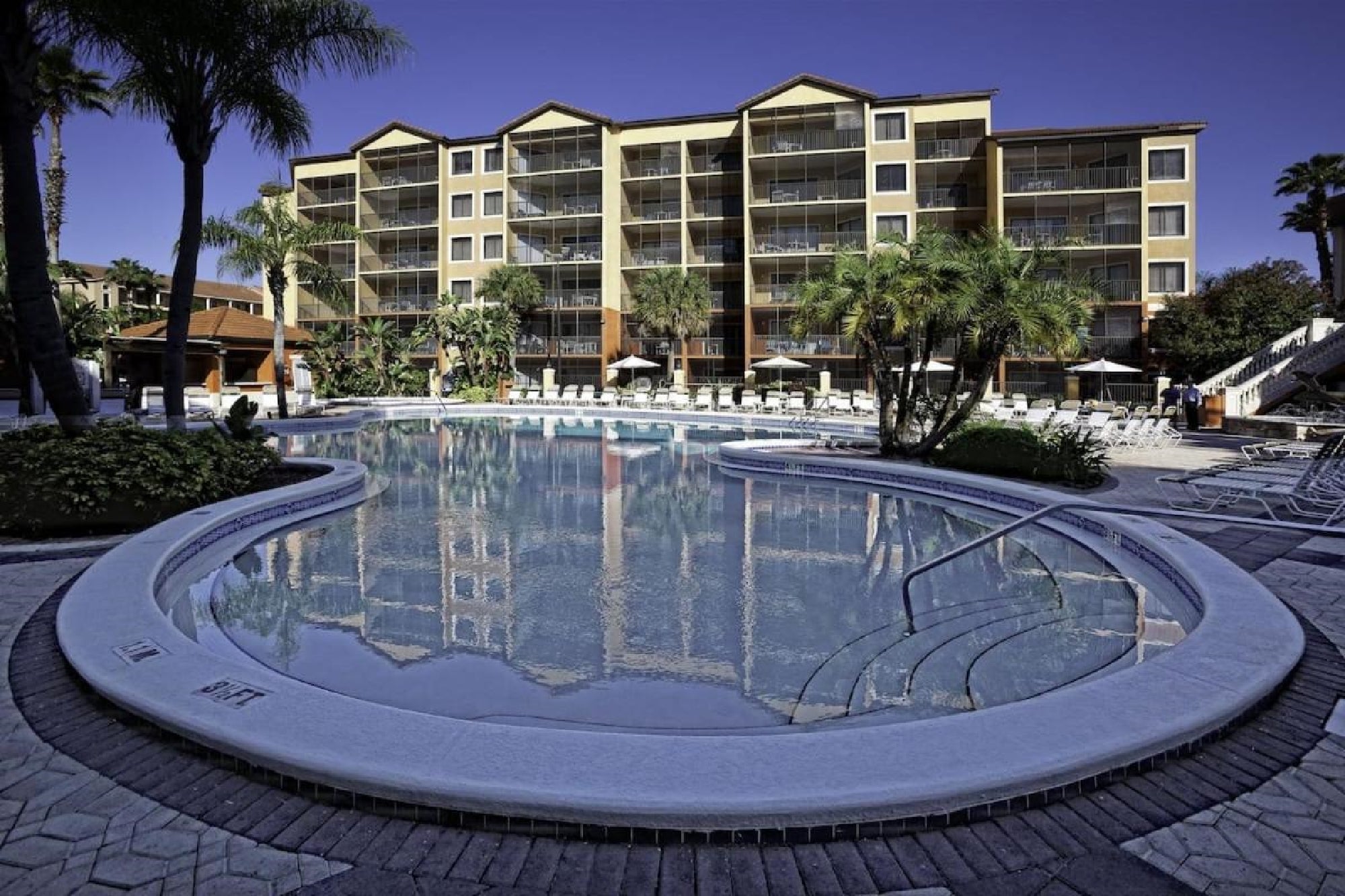 Outdoor hotel pool with surrounding palm trees, lounge chairs, and a multi-story building in the background under a clear blue sky.