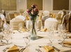 A round table is set for an event with a floral centerpiece, neatly arranged silverware, wine glasses, and yellow napkins. Chairs are covered and tied with gold bows.