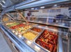 Buffet counter with trays of assorted food, including sausages, pasta, vegetables, and more, under a glass cover.