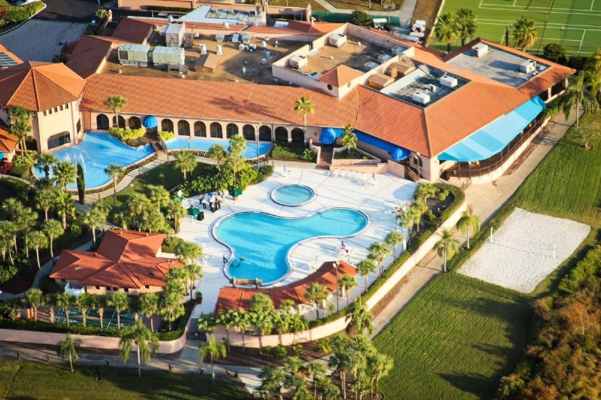 Aerial view of a large resort complex with multiple pools, palm trees, and a circular building. Tennis courts are visible in the background.