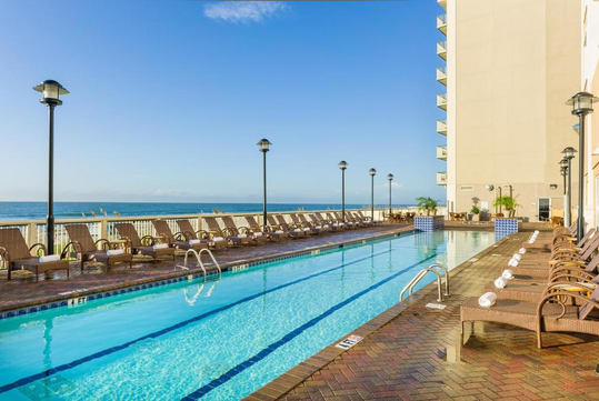Rectangular outdoor swimming pool with lounge chairs beside it, overlooking the ocean, next to a tall building under a clear blue sky.
