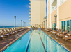 Rectangular outdoor pool beside a tall hotel building, with lounge chairs and towels arranged along both sides, overlooking the ocean under a clear blue sky.