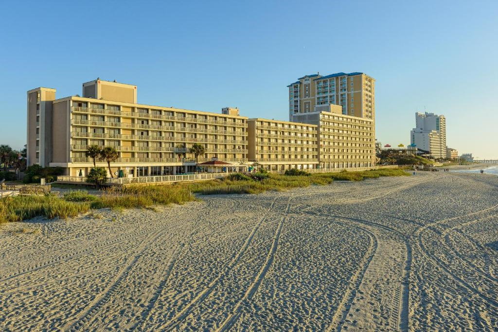 Wide sandy beach with tire tracks in the foreground, several multi-story beachfront hotels and palm trees in the background under a clear blue sky.