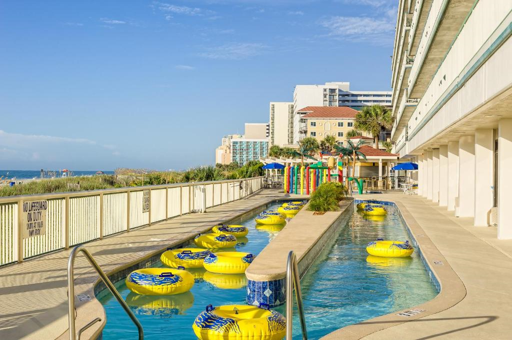 A hotel outdoor lazy river with yellow inflatable tubes, next to a multi-story building; the beach and other hotels are visible in the background.
