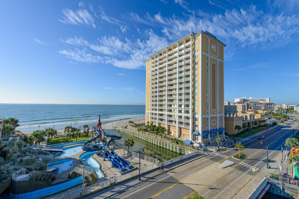 A tall yellow beachfront hotel stands beside a water park with slides, overlooking the ocean under a blue sky with scattered clouds.