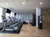 A modern gym with several elliptical machines, a multi-station weight machine, two TVs, a water dispenser, and a towel rack, all on tiled flooring with large windows.