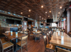 Spacious restaurant interior with wooden tables and chairs, decorative ceiling panels, multiple TVs, framed wall art, and a mix of stone and brick walls.
