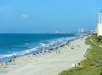 Wide sandy beach with people, umbrellas, and gentle waves along a shoreline. Tall buildings and green vegetation are visible to the right under a clear blue sky.