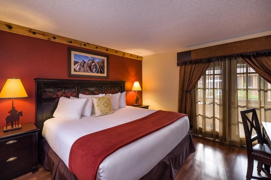 A hotel room with a king-size bed, red accent wall, two lamps, western-themed decor, a painting above the bed, and a window with sheer curtains letting in daylight.