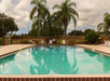 Outdoor swimming pool with clear water, surrounded by a concrete deck, palm trees, lounge chairs, and a wooden fence under a cloudy sky.