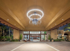 A well-lit hotel entrance with a large circular chandelier, flanked by palm trees and glass doors under a brown ceiling.
