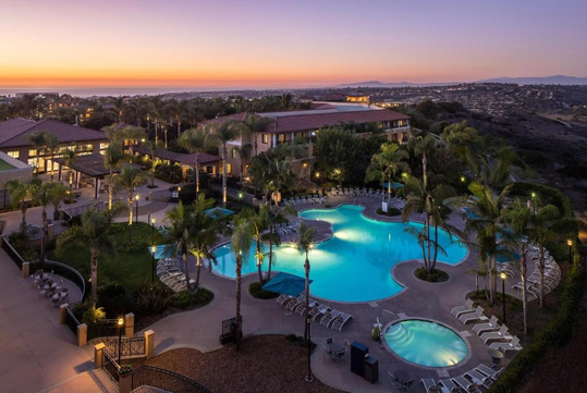 Aerial view of a resort at sunset featuring a large, uniquely shaped pool, surrounded by palm trees and lounge chairs, with a scenic landscape in the background.