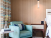 A cozy corner with a teal armchair, striped pillow, wooden side table, decorative vase, and hanging light, set against a wood-patterned wall and patterned curtains.