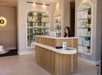 Reception area with a wooden desk, a computer screen, and a smiling receptionist. Behind are shelves displaying various products and decor items under soft lighting.