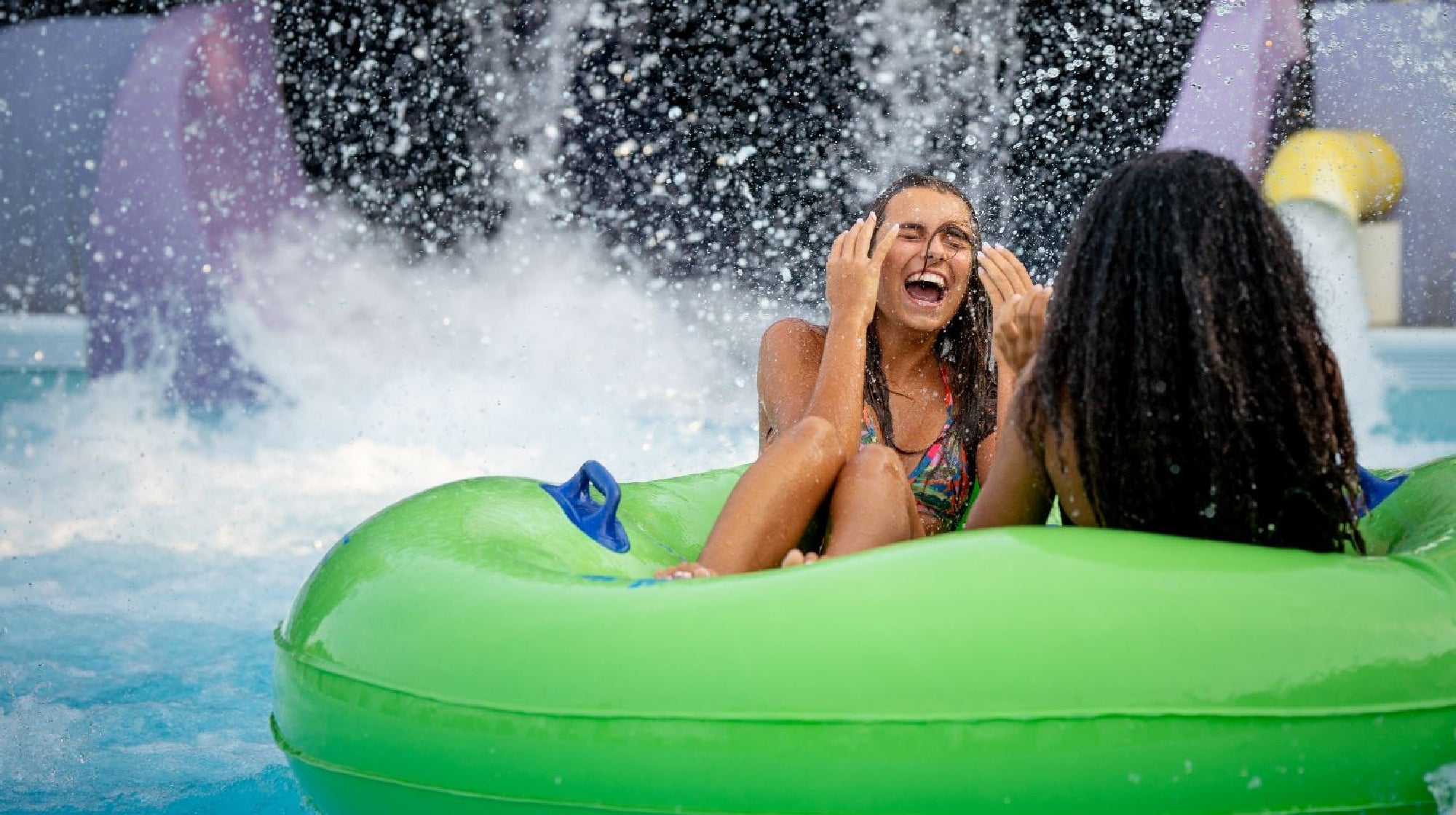 Two people in swimsuits sit on a green inflatable tube under splashing water at a water park, laughing and enjoying the ride.