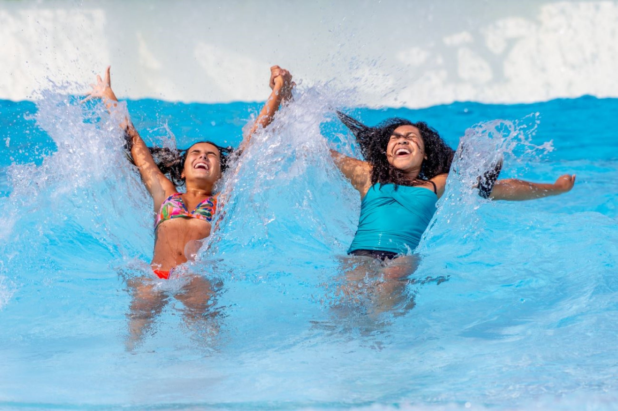 Two women in swimsuits splash water as they lean back in a pool, smiling and lifting their arms with water spraying around them.