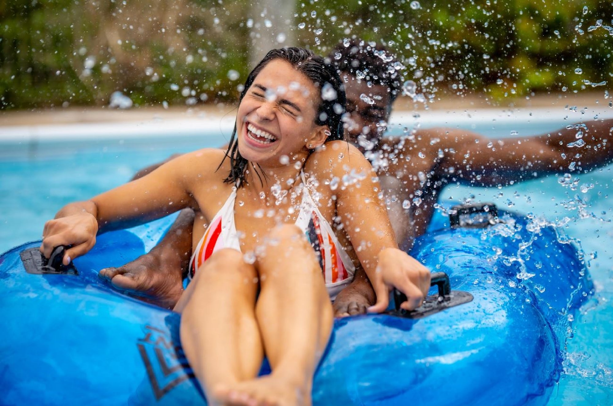 Two people ride a blue inflatable tube in a pool, laughing and smiling as water splashes around them.