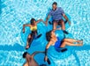 Four people float in a pool on blue inner tubes, holding hands and smiling at each other under bright sunlight.