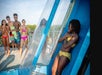 A girl in a yellow swimsuit stands inside a transparent water slide capsule, while four other teens in swimsuits watch and smile nearby.