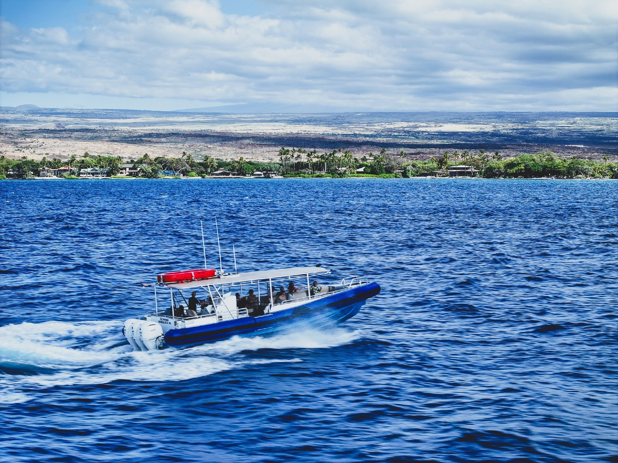 A small motorboat travels quickly across blue ocean water, with a distant coastline and cloudy sky in the background.