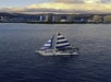 A sailboat with blue and white striped sails is on the water near a city skyline with tall buildings and mountains in the background.