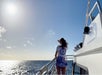 A woman stands on the deck of a boat, facing the ocean and the sun, with clear skies and calm water in the background.