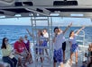A group of people on a boat; three crew members are singing and dancing while passengers watch and sit around the deck. Ocean and distant land visible in the background.
