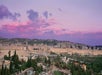A panoramic view of Jerusalem’s Old City walls at sunset, with historic buildings, greenery, and parked cars in the foreground under a purple-pink sky.