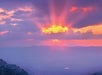 Vibrant sunset with sun rays shining through clouds over a hazy, mountainous landscape.