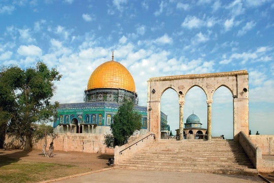 The Dome of the Rock with its golden dome sits beside a stone archway and stairs under a partly cloudy sky.