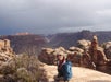 A woman wearing sunglasses and a jacket posing at the top of the rock formation at the Canyonlands with the White Rim Trail in the background.