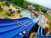People slide down a multi-lane water slide on mats at a water park. Several slides and a pool are visible, with trees and park buildings in the background.