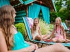 Three people by a poolside; an adult in the foreground and two children sitting on loungers, smiling and chatting.