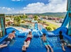 Four people lie on mats at the top of a water slide, preparing to race. The slide overlooks a water park and town, with mountains in the background under a blue sky with clouds.