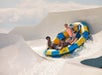Four people ride a yellow and blue raft down a water slide under a sunny sky.