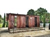 A rusted metal jail cell structure with open barred doors stands on a concrete platform surrounded by trees and greenery.