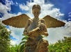 A bronze angel statue with large wings holds a baby in its arms, set against a partly cloudy sky and surrounded by greenery.