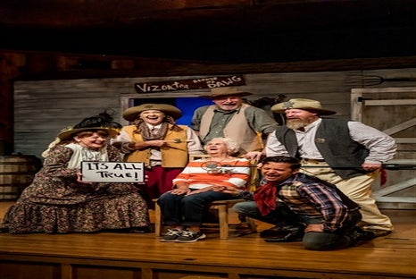 Six people in Western costumes pose on stage; one holds a sign reading "IT'S ALL TRUE!" and another sits in a chair looking surprised. A western saloon set is in the background.