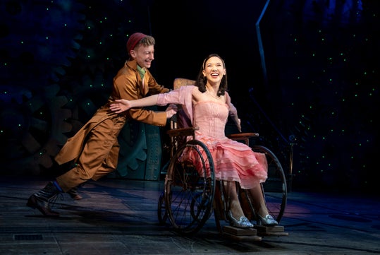 Two performers are on stage. One is pushing the other, who is sitting in a wheelchair and smiling, both in costume with a background of gears and green lights.