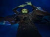 A person dressed as Elphaba from "Wicked" stands with green skin, wearing a dark, flowing dress, and holding arms raised against a blue-lit background.