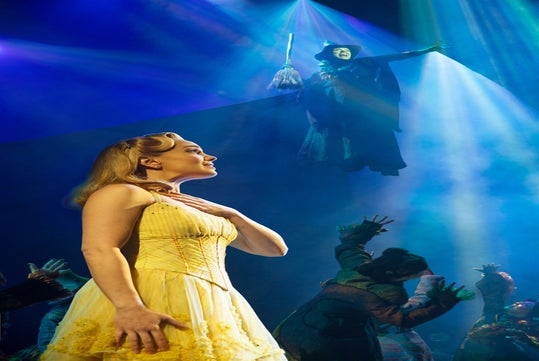 A woman in a yellow dress stands on stage with a hand on her chest, while another figure with a broom is elevated above her in dramatic blue and green lighting.