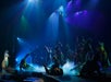 A theatrical stage performance with actors in dynamic poses, mist effects, and dramatic lighting highlighting a figure suspended in the air above the ensemble.