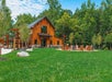 A wooden lodge with a patio and Adirondack chairs sits on a green lawn, surrounded by trees and under a partly cloudy sky.