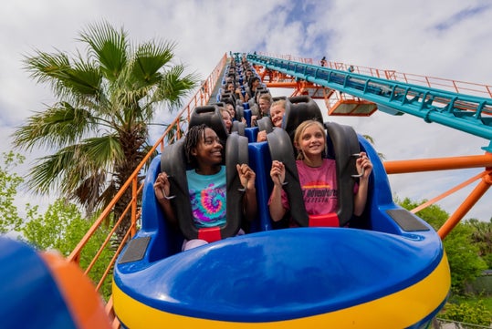 People sit in the front row of a roller coaster car, holding onto the safety bars as the ride ascends a steep track, with palm trees and a partly cloudy sky in the background.