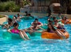 People float on inflatable tubes in a lazy river at a water park, with two women in the foreground reaching out to hold hands.