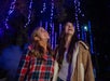 Two young women stand outdoors at night, looking up and smiling, with blue string lights and trees in the background.